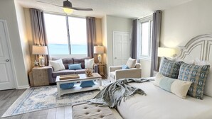 Un dormitorio amplio con una cama grande, un sofá, una silla y una mesita de noche. Se ve el océano a través de las ventanas.