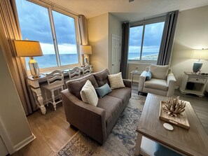 A living room with a brown sofa, two armchairs, a coffee table, and a view of the ocean through large windows.