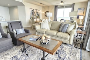 Una sala moderna con un sofá color beige, un sillón oscuro y una mesa de centro con objetos decorativos. Hay un comedor con una mesa y sillas, y un ventanal que ofrece una vista.