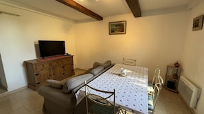 A room with a sofa, a dining table with a white tablecloth, a television on a wooden cabinet, and a painting on the wall. - Paul à 12 km de la mer (ROQUEBRUNE SUR ARGENS)
