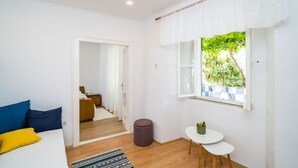 A bedroom with a bed, a small table, a window with a view of greenery, and a door leading to another room. - Apartment Marija 5 mins away from Old Town (Dubrovnik)