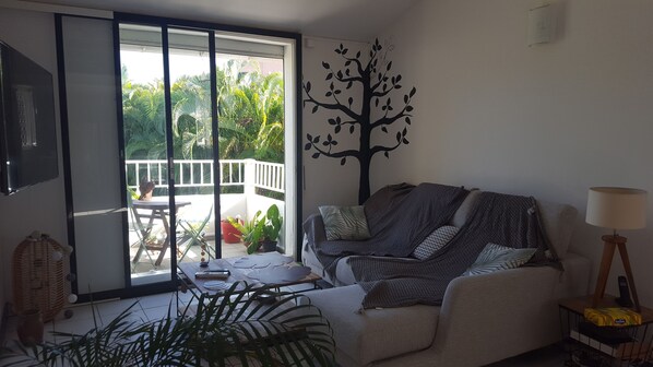 Un salon comprenant un canapé, une lampe, un motif décoratif représentant un arbre appliqué sur le mur, et une vue sur un balcon agrémenté de plantes.