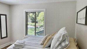 Een slaapkamer met een bed, een raam met zonwering, een ingelijst beeld aan de muur en een stap opgevouwen handdoeken.