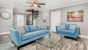A modern living room with a blue sofa, a glass coffee table, a ceiling fan, and a staircase.