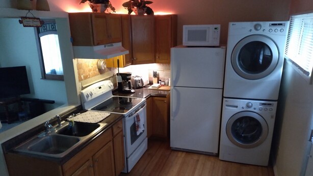 A compact kitchen with wooden cabinets, a microwave, a refrigerator, a stove, and a washing machine. - Cozy Up-North Townhouse Bungalow (harbor springs)