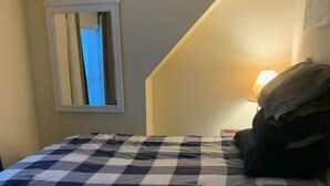 A bedroom with a checkered bedspread, a window with a view of a building, and a lamp on the nightstand. - Cozy Up-North Townhouse Bungalow (harbor springs)