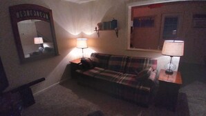 A living room with a plaid sofa, two lamps, a mirror, and a window with blinds. - Cozy Up-North Townhouse Bungalow (harbor springs)