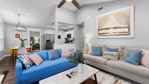 A modern living room with a blue sofa, a grey sofa, and a coffee table. There is a ceiling fan, a chandelier, and a painting on the wall. - 4BDR+Pets OK+Huge Yard+FirePit+3mintoBeach (Rockport)