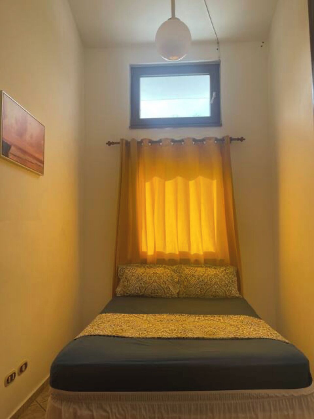 Una habitación pequeña y acogedora con cama, ventana con cortinas e imagen colgada en la pared.