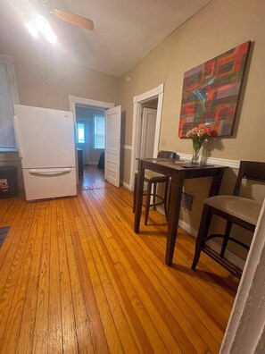 Une cuisine avec un plancher en bois, un réfrigérateur, une table à manger avec des chaises et un tableau accroché au mur.