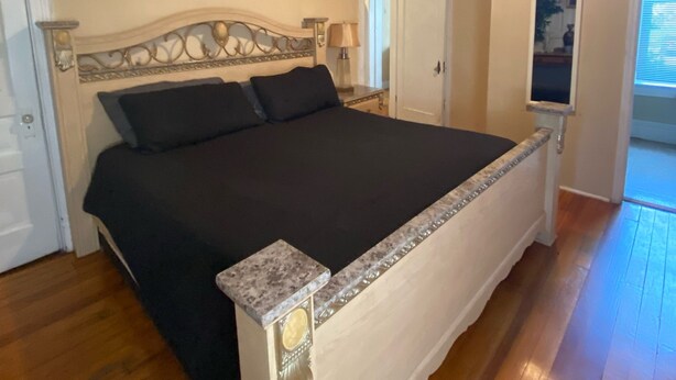 A bedroom with a large bed, a headboard, a painting on the wall, and a window with blinds. - Pet Friendly First Floor 2 Bedroom Apartment (St. Louis)