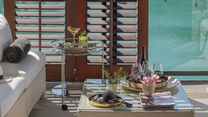 A sunlit outdoor seating area with a glass table, cushions, and a view of a pool.