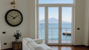 Un salon avec un canapé, une horloge murale, une plante en pot et une vue sur la mer par une porte vitrée.