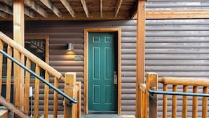 Una cabaña de madera con puerta verde, escaleras de madera y barandilla metálica.