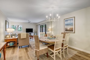 A dining area with a round table, white chairs, a chandelier, and a painting on the wall.
