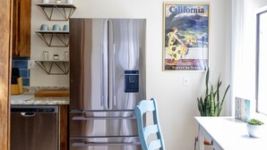 A modern kitchen with a stainless steel refrigerator, a dishwasher, and a dining area with a blue chair and a white table. - Sea La Vie - At the entrance to MacKerricher State Park and is in walking distance (Fort Bragg)