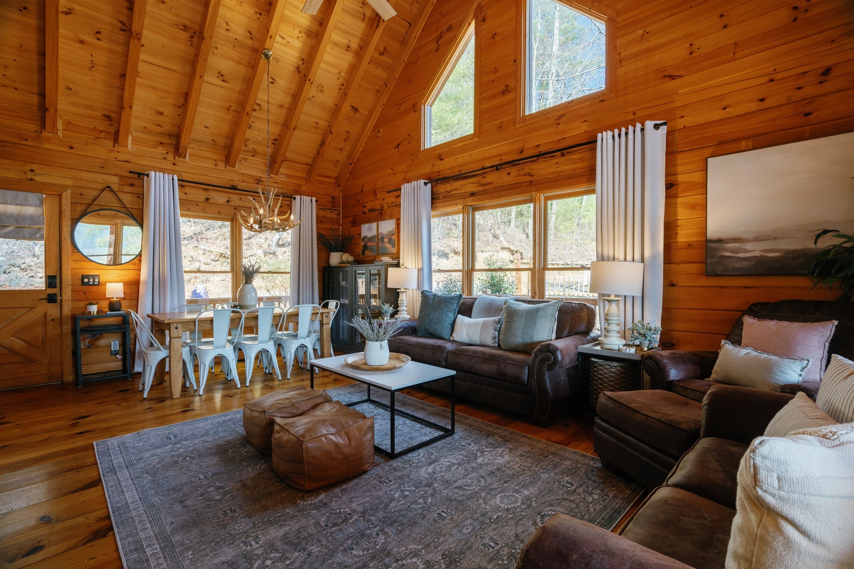 Een gezellig cabin interieur met houten wanden, een eethoek met witte stoelen, een bruine leren bank en een groot raam met uitzicht op de natuur.