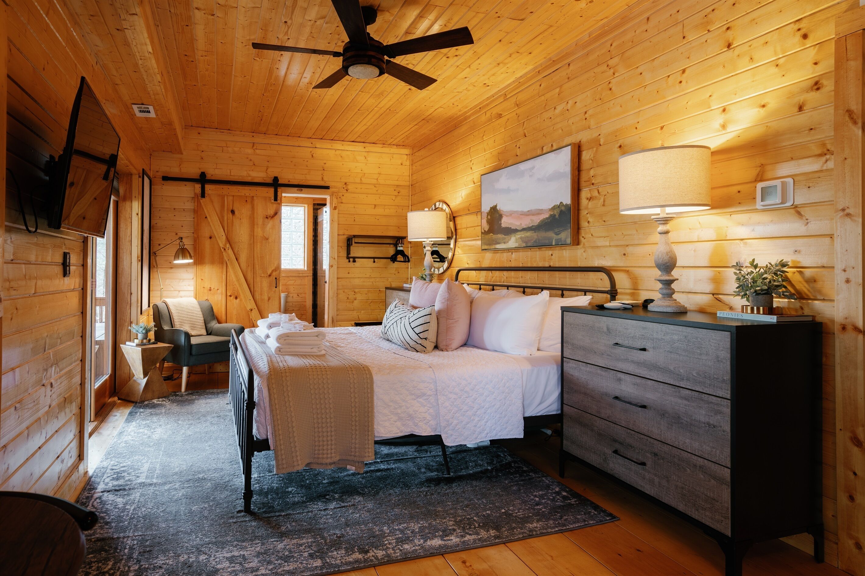 A wooden cabin bedroom with a bed, bedside table, lamp, and a painting on the wall.