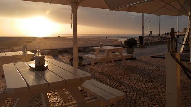An outdoor seating area with tables and benches under a canopy, overlooking a beach with people in the distance. - 2 Bedroom Apartment a Walk from the Beach of Praia da Areia Branca. (Lourinhã)