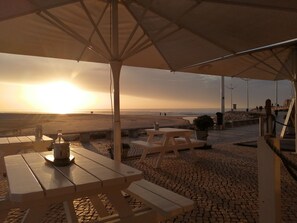 An outdoor seating area with tables and benches under a canopy, overlooking a beach with people in the distance.