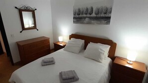 A neatly made bed with pillows and folded towels, a wooden dresser, a bedside table with a lamp, a wall-mounted mirror, and a painting of trees. - 2 Bedroom Apartment a Walk from the Beach of Praia da Areia Branca. (Lourinhã)