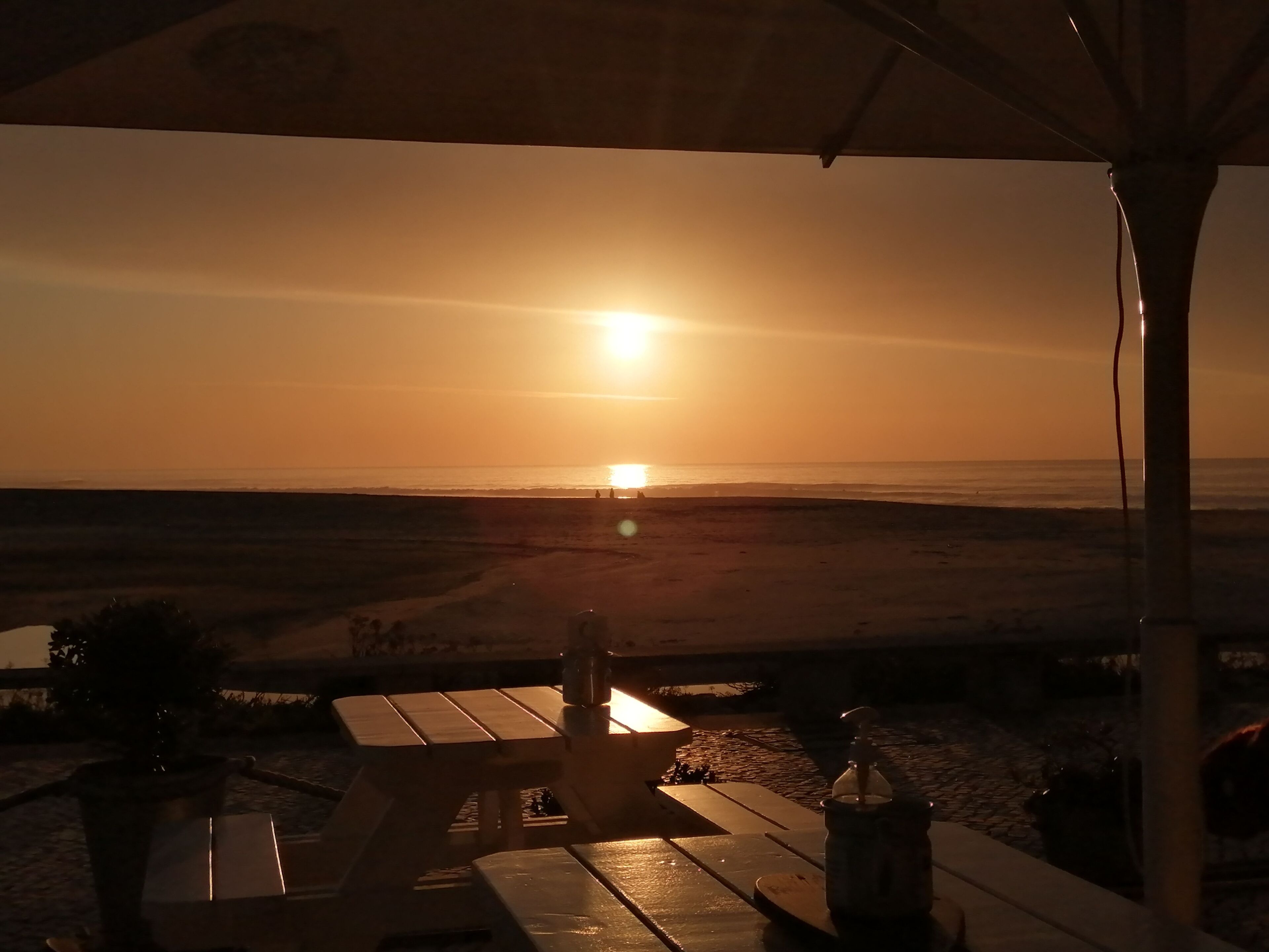 Vista del atardecer desde un patio frente a la playa, con mesas y sillas de madera, una planta en maceta y una botella de agua.