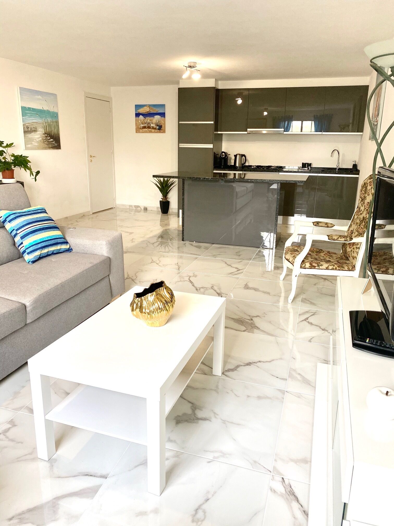 Un salon moderne avec un canapé gris, une table basse blanche et un fauteuil à motifs. On aperçoit, au fond, une cuisine avec des meubles sombres et un îlot central.