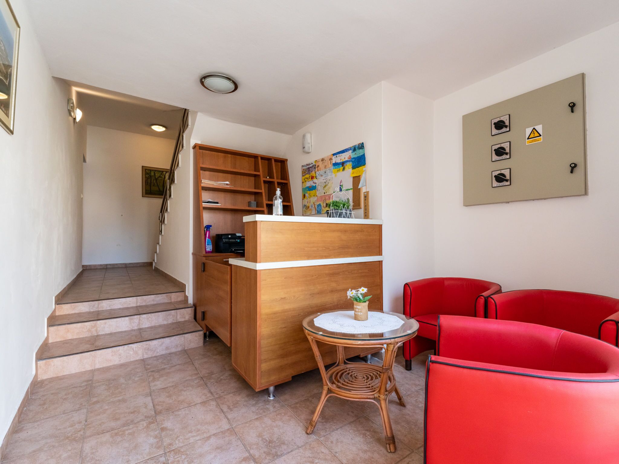 Eine Hotelhalle mit einem hölzernen Empfangstresen, einem kleinen runden Tisch mit einer Vase, roten Stühlen, einer Treppe und einem Bulletinbrett mit Sicherheitshinweisen.