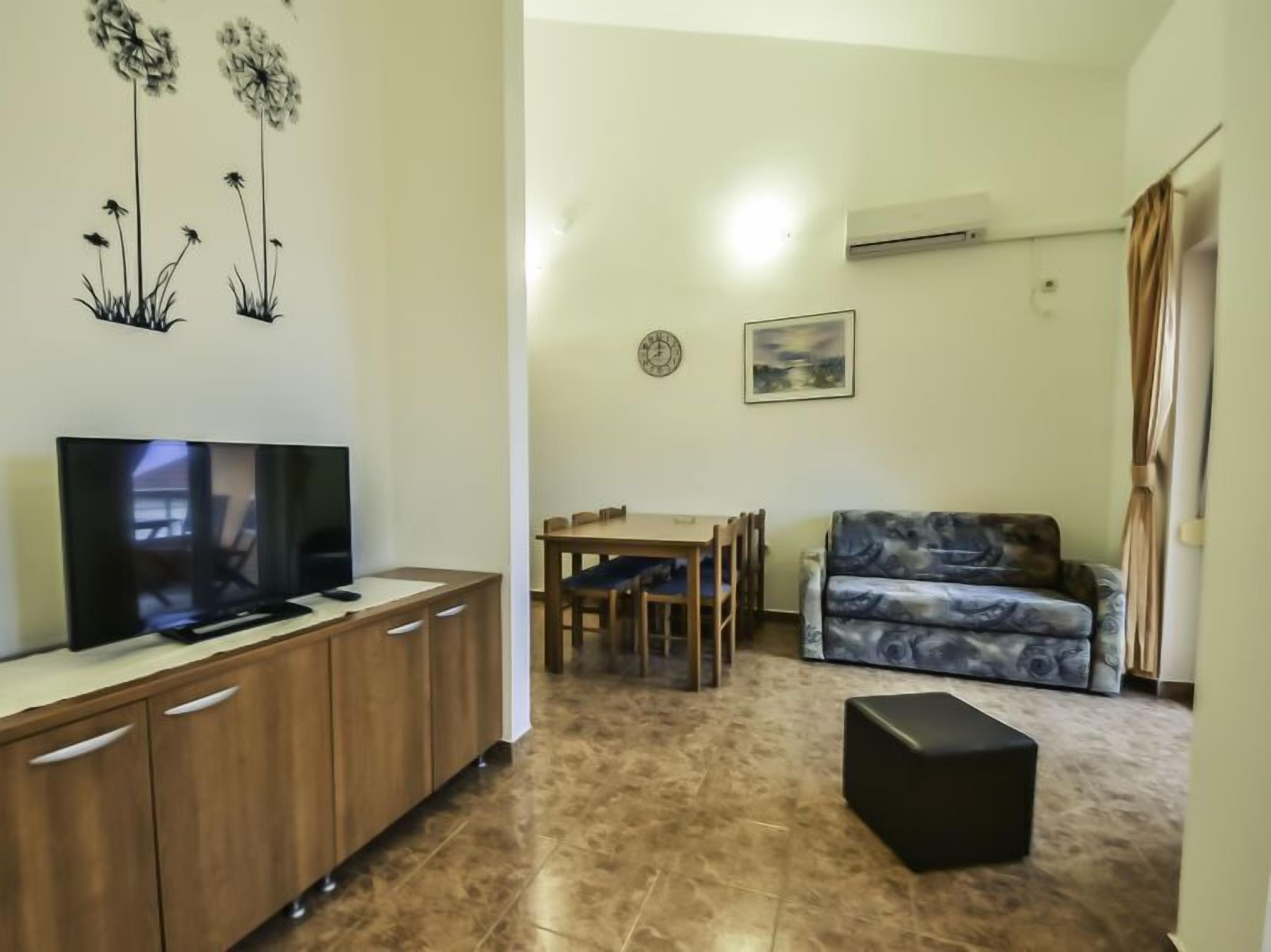 A living room with a TV, wooden cabinet, dining table, chairs, sofa, and a black ottoman.
