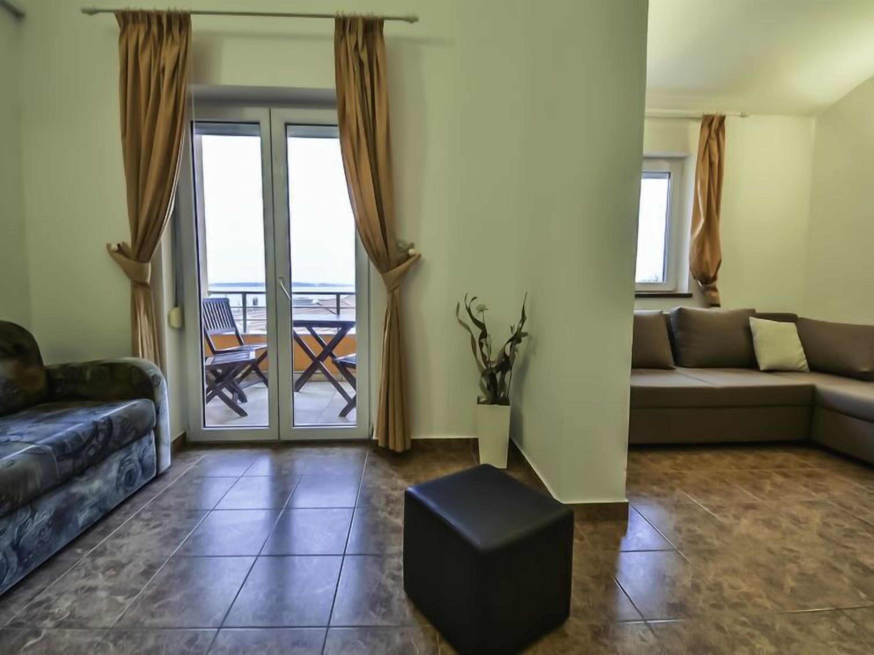 A living room with a sofa, a patterned armchair, a black ottoman, and a sliding glass door leading to an outdoor area with a table and chairs.