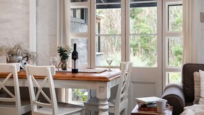 Un espace repas avec une table en bois, des chaises blanches et une vue sur la verdure grâce à de grandes fenêtres.