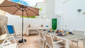 Eine Terrasse mit Esstisch, umgeben von weißen Stühlen, einem großen Sonnenschirm und einem grünen Tor.
