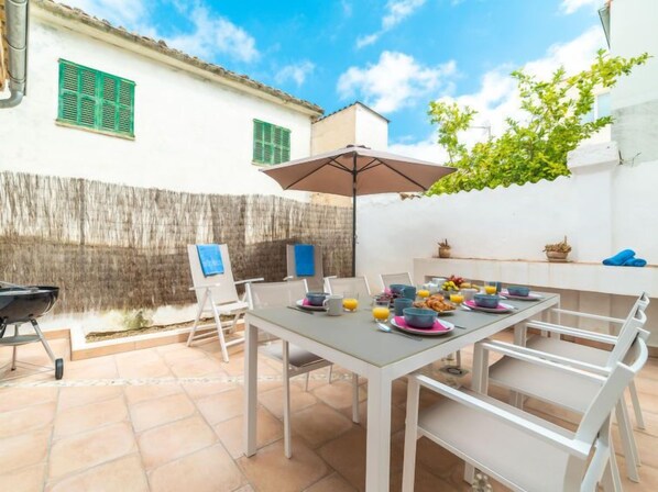 An outdoor patio with a table set for breakfast, chairs, a grill, and a view of a white building with green shutters. - Can Monjo by Interhome (Andratx)