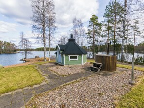 A small cabin with a green roof and a chimney, situated on a gravel path with a lake in the background.