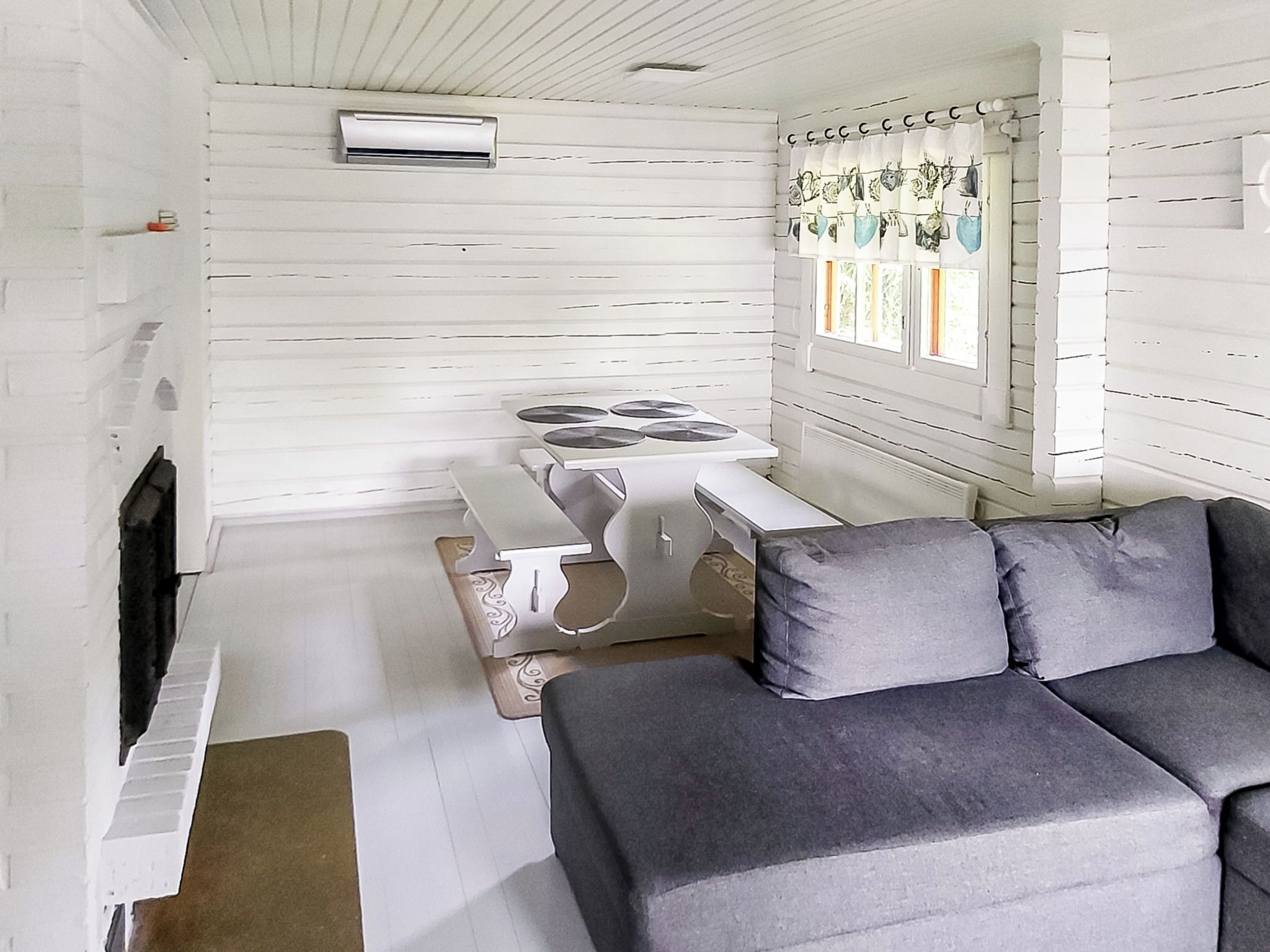 Una habitación con una pared de madera blanca, una mesa blanca con una silla blanca, un sofá gris y una ventana con cortinas.