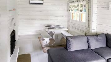 Um quarto com parede de madeira branca, uma mesa branca com uma cadeira branca, um sofá cinzento e uma janela com cortinas.