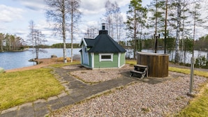 A small cabin with a green exterior and a black roof, situated on a gravel path with a wooden deck and a hot tub nearby.