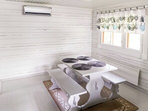 A room with white wooden walls, a white table with four circular objects, a window with white curtains, and an air conditioning unit. - Villa koivumäki by Interhome (Kankaanpää)
