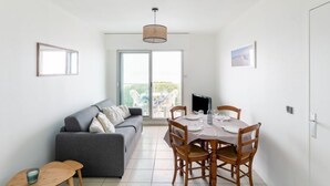 A modern living room with a grey sofa, a dining table set for four, and a large window with a view of greenery. - Apartment Claire by Interhome (Arcachon)