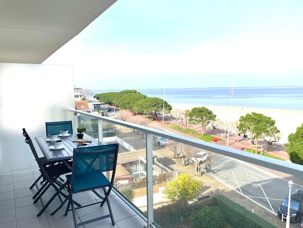 A balcony with a table and chairs, overlooking a beach and parking area.