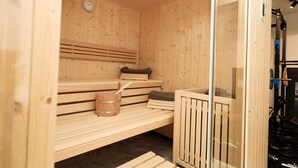 Sauna de fusta amb bancs, un cubet de fusta i una galleda, i una porta de vidre que dona a una zona de gimnàs.