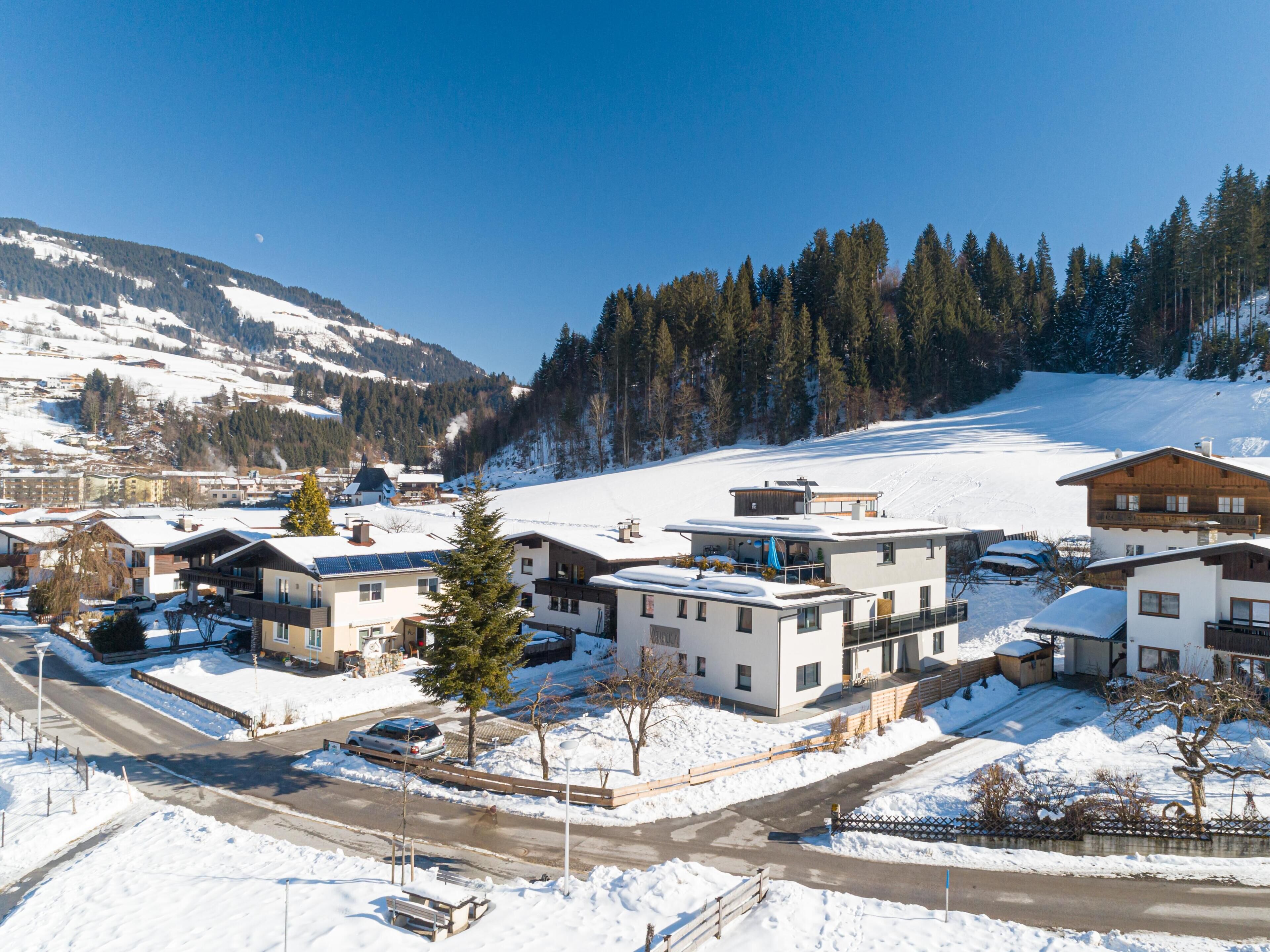 Ein verschneiter Wohnbereich mit modernen Häusern, einer Straße und einem bewaldeten Berg im Hintergrund.