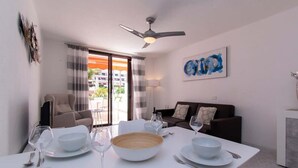 A dining area with a white table set for four, a ceiling fan, and a view of a balcony with a building in the background. - Parque Santiago II 131 - One Bed Duplex with Pool (Playa de las Americas)