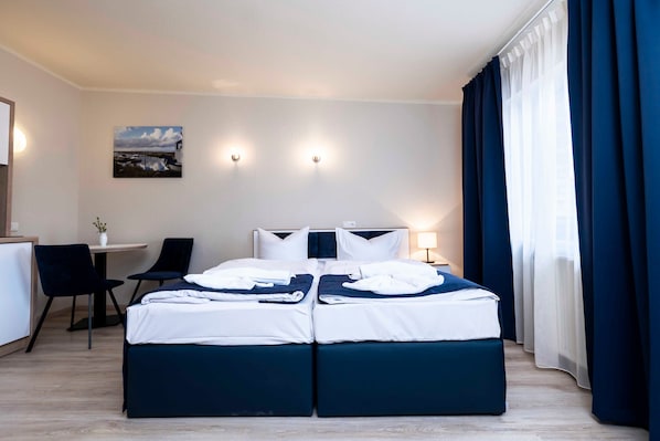 A hotel room with a double bed, a desk with a chair, a small table, and a painting on the wall.