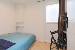 A small, sparsely furnished room with a bed, a chair, and a window.
