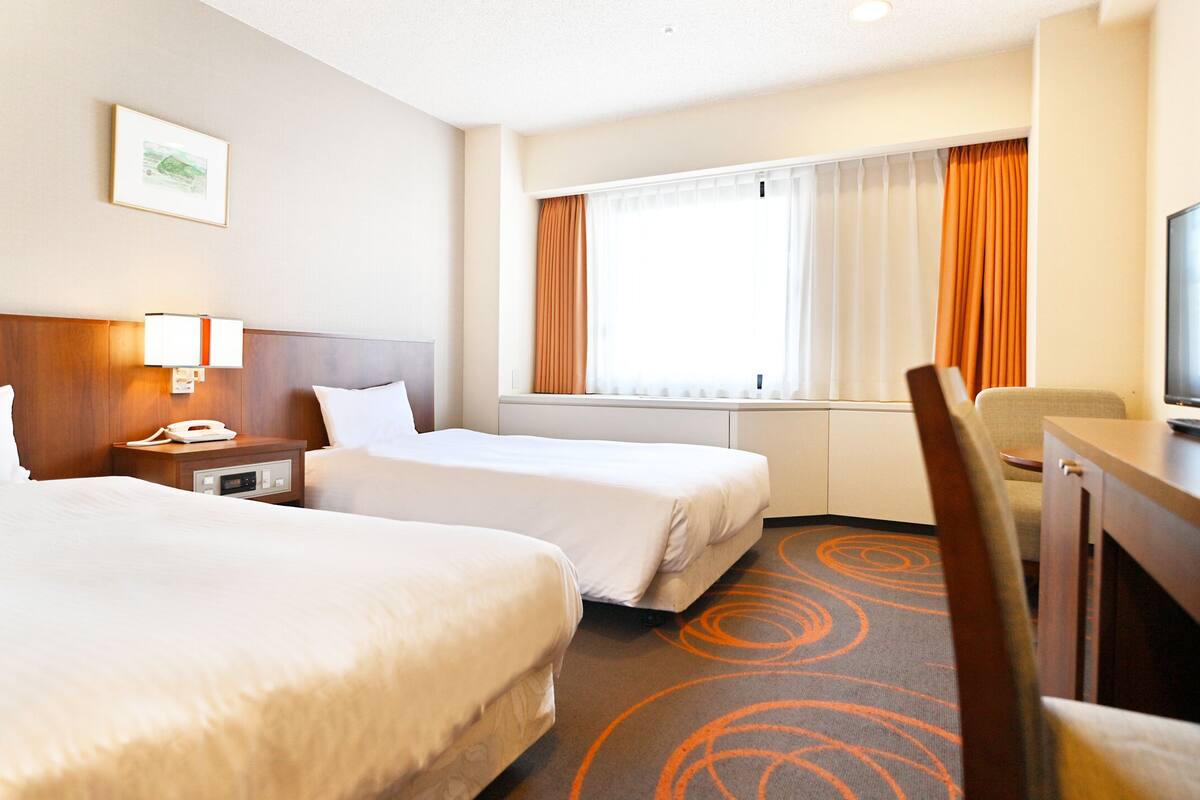 A hotel room with two beds, a desk, a TV, a window with curtains, and a framed picture on the wall.