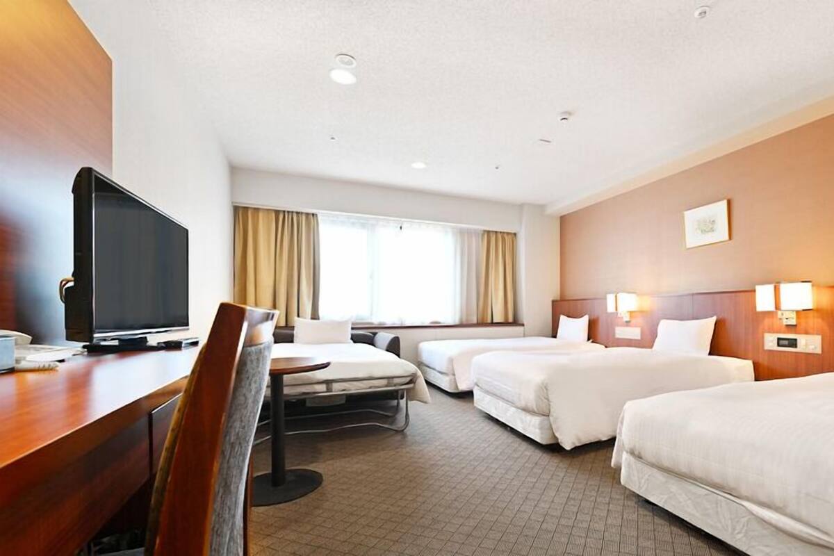 A hotel room with a desk, TV, two beds, and a window with curtains.