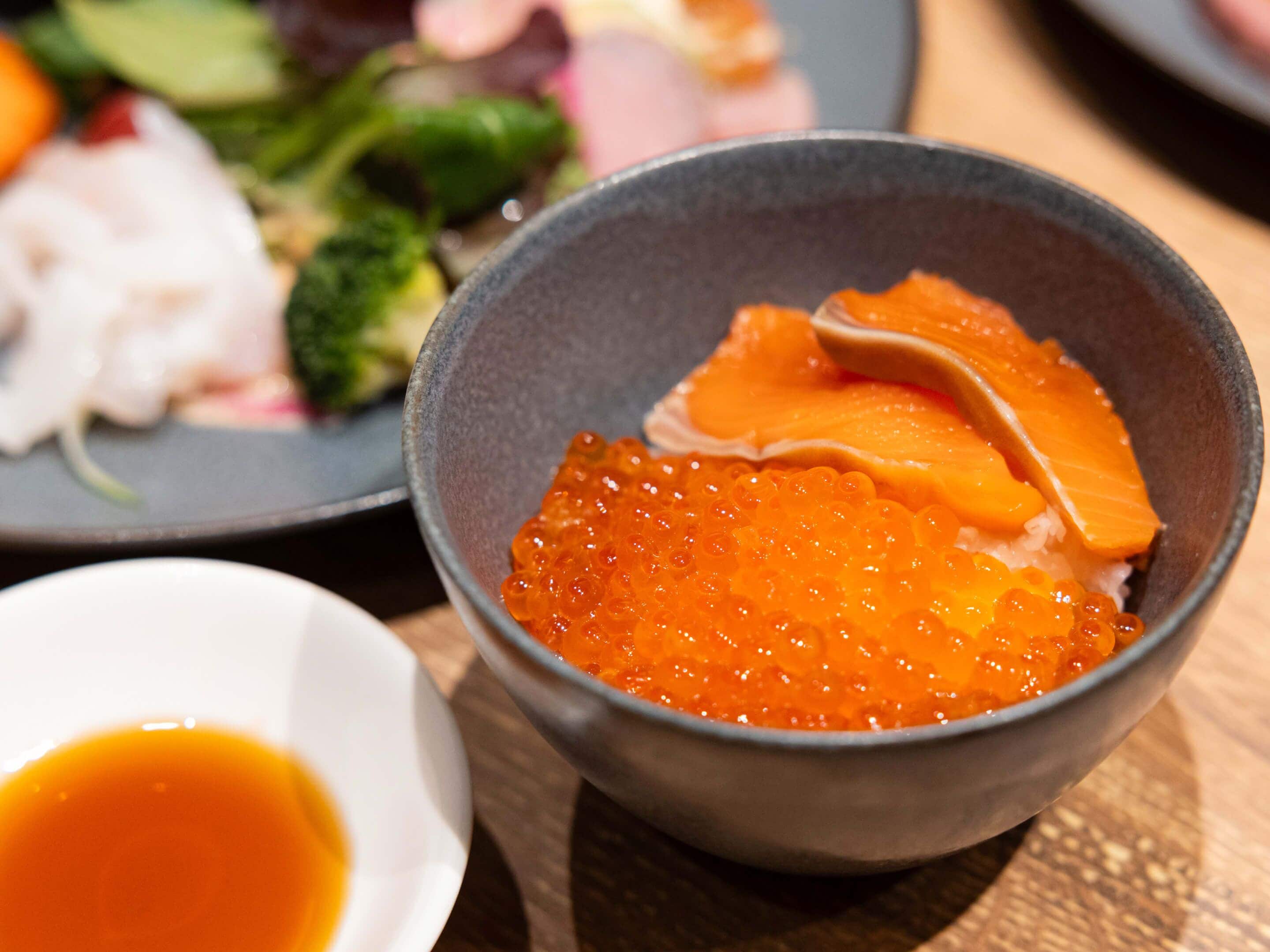 A bowl of salmon roe on rice, a small bowl of soy sauce, and a plate with various dishes including vegetables and fish.