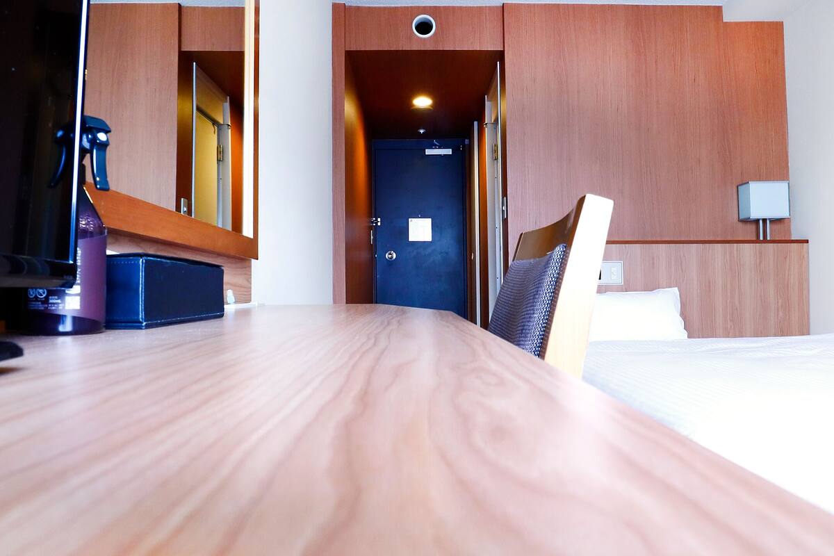 A hotel room with a wooden desk, a bed, a chair, a nightstand, and a lamp.