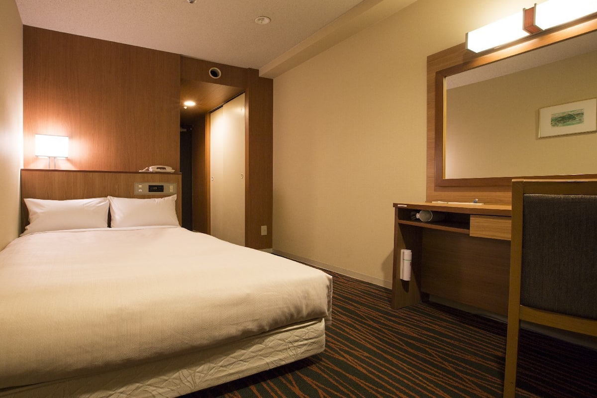 Una habitación de hotel con cama, escritorio, silla, espejo y teléfono en la pared.
